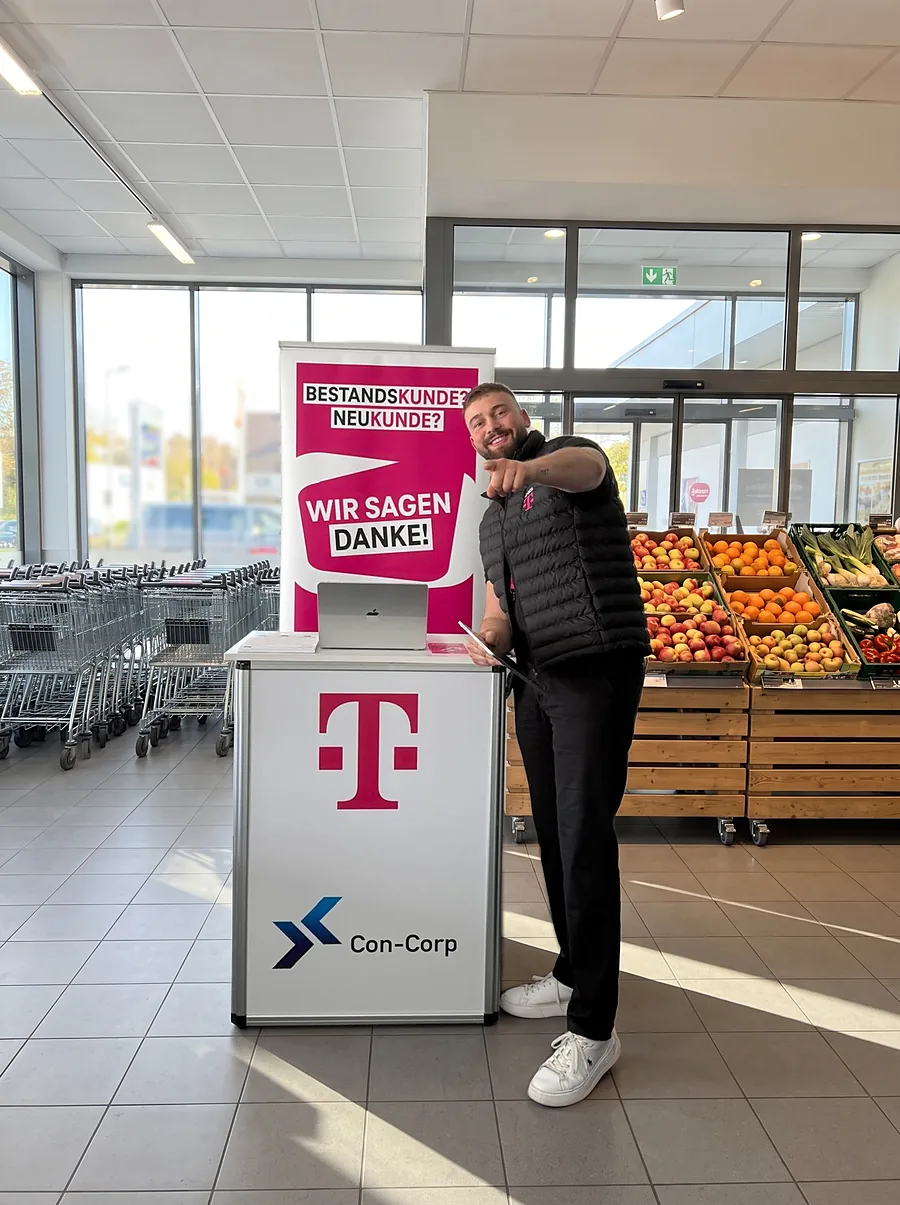 Promoter zeigt lachend in die Kamera, neben ihm unser Stand mit Telekom und Con-Corp Logo
